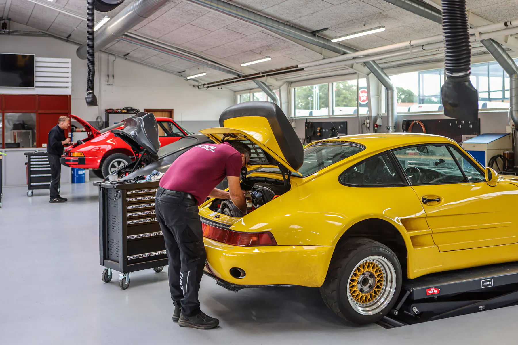 Mekaniker arbejder i motorrummet på en gul klassisk bil i Classikos værksted